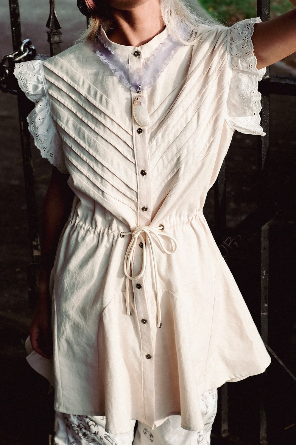 Person wearing a light-colored outfit with a textured pattern, standing against a dark metal gate.