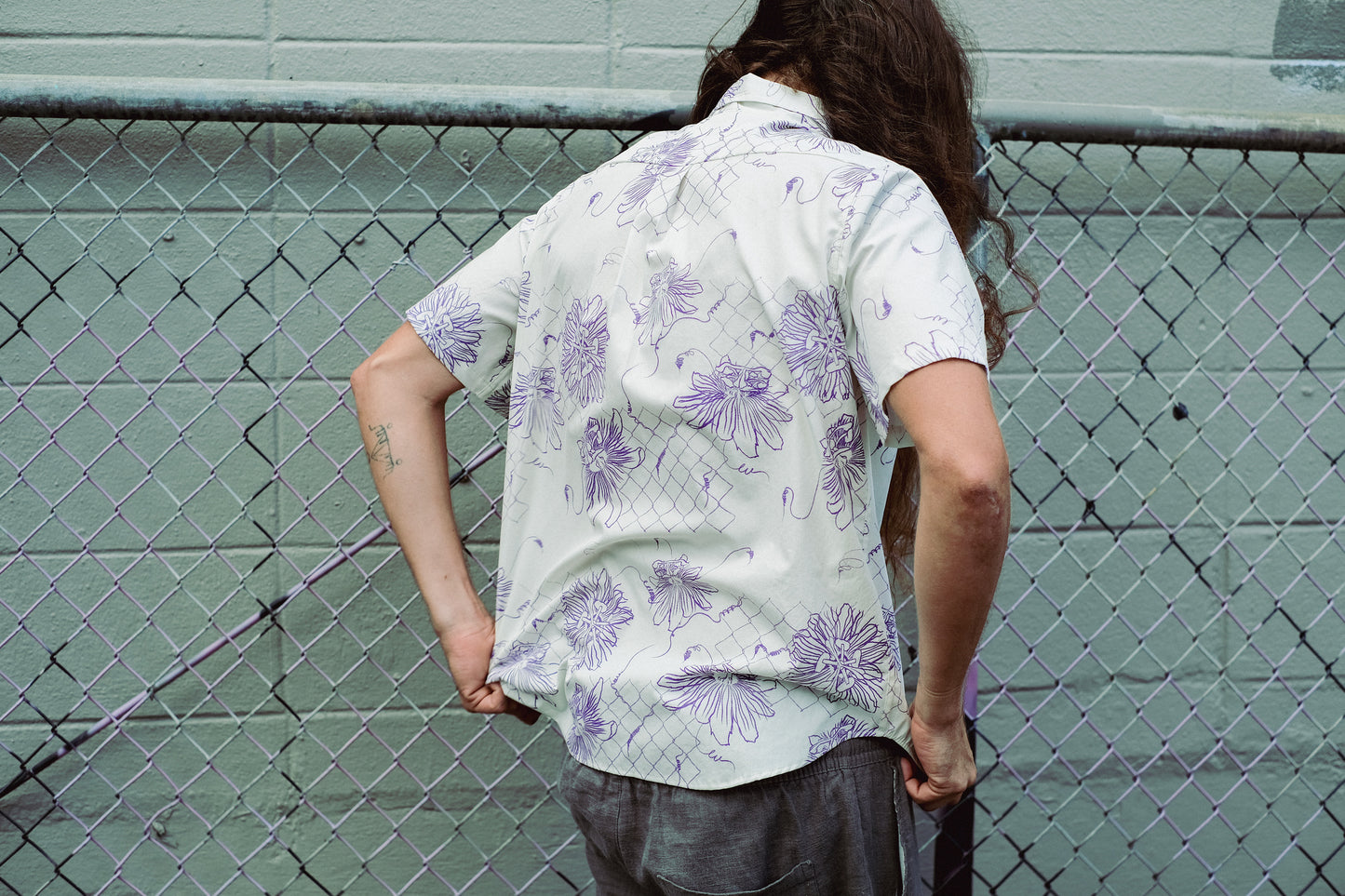 Person wearing a white shirt with purple floral patterns against a chain-link fence.